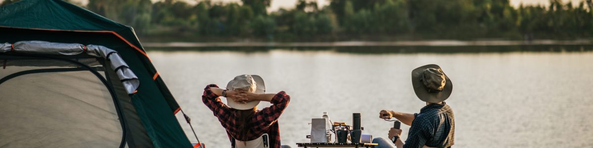 Rear view of young backpacker couple sitting to relax at front of the tent near the lake with coffee set and making fresh coffee grinder while camping trip on summer vacation
